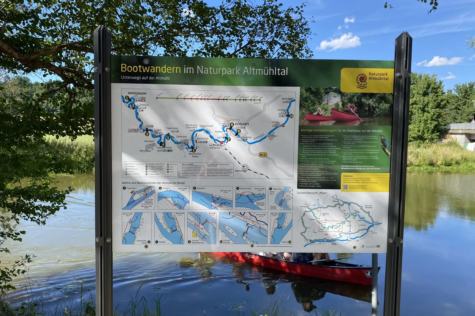 Infotafel „Bootwandern im Naturpark Altmühltal“ mit Flusskarte am Ufer, dahinter ein rotes Kanu